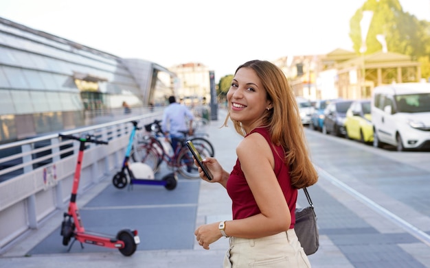 Une jeune femme d'affaires confiante se retourne en regardant la caméra et en souriant à l'aide d'une application pour smartphone, elle se prépare à utiliser un scooter électrique pour rentrer chez elle