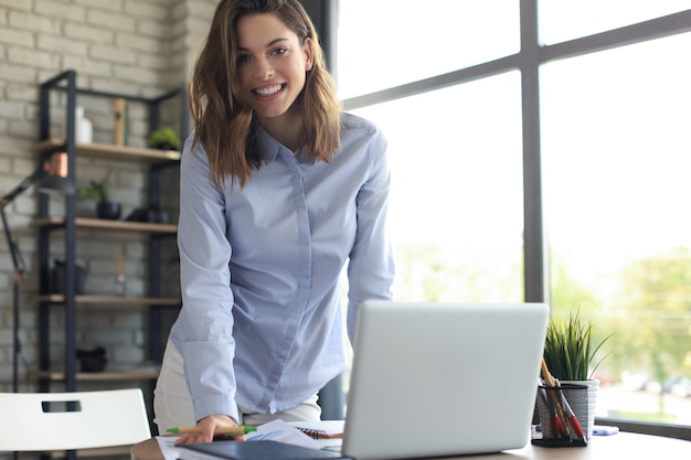 Jeune femme d'affaires confiante avec un sourire amical, debout derrière son bureau dans un bureau à domicile en regardant la caméra.