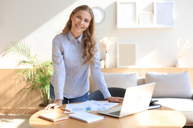 Jeune femme d'affaires confiante avec un sourire amical, debout derrière son bureau dans un bureau à domicile.