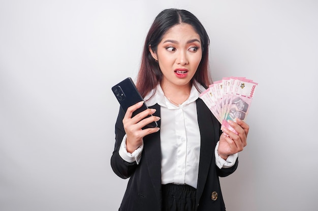 Une jeune femme d'affaires confuse porte un costume noir tenant son téléphone et de l'argent en roupie indonésienne isolé par un fond blanc