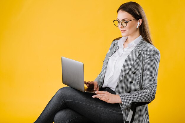 Jeune femme d'affaires en costume portant des lunettes assis et utilisant un ordinateur portable sur fond jaune