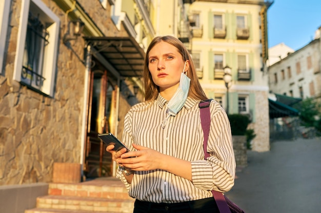 Jeune femme d'affaires employée de bureau en masque de protection médicale avec smartphone dans les mains masque sur l'oreille visage ouvert Femme dans la rue de la ville Entreprise de style de vie dans une épidémie de pandémie mauvaise écologie