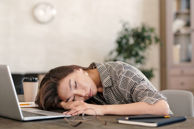 Jeune femme d'affaires épuisée en tenue de soirée gardant la tête à portée de main tout en faisant la sieste devant un ordinateur portable après le travail dans un environnement de bureau