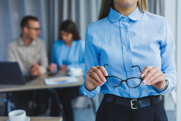 Jeune femme d'affaires Gestionnaire femme dans des verres classiques souriant pendant le temps de travail au bureau avec des collègues d'ordinateur portable en arrière-plan Un collègue est en arrière-plan mise au point sélective