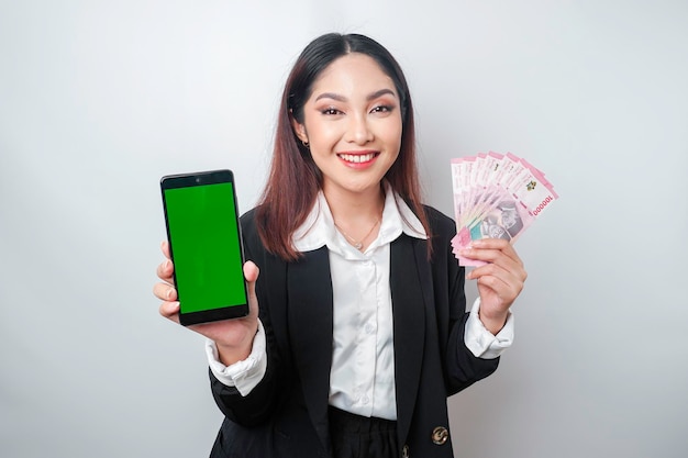 Une jeune femme d'affaires heureuse porte un costume noir montrant son téléphone et de l'argent en roupie indonésienne isolé sur fond blanc