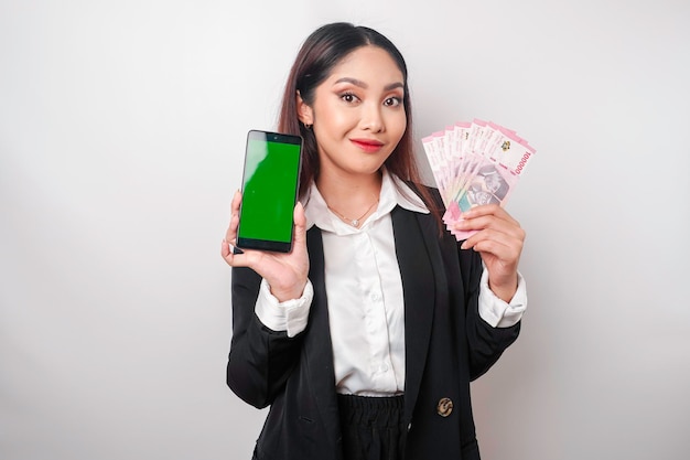 Une jeune femme d'affaires heureuse porte un costume noir montrant son téléphone et de l'argent en roupie indonésienne isolé sur fond blanc