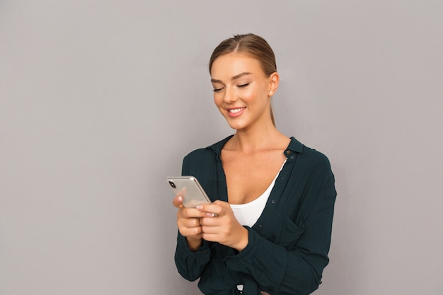 Jeune femme d'affaires heureuse posant isolée sur fond de mur gris à l'aide de téléphone portable.