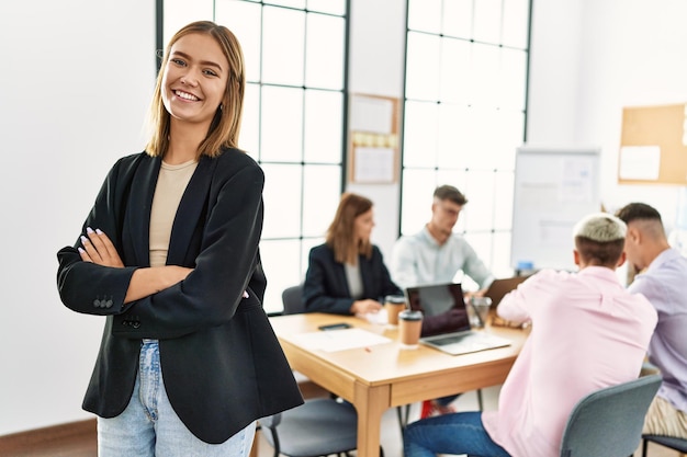 Jeune femme d'affaires hispanique souriante heureuse debout avec les bras croisés au bureau lors d'une réunion d'affaires