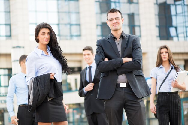 Jeune femme d'affaires et homme d'affaires avec son équipe en arrière-plan debout devant l'immeuble de bureaux, regardant la caméra.