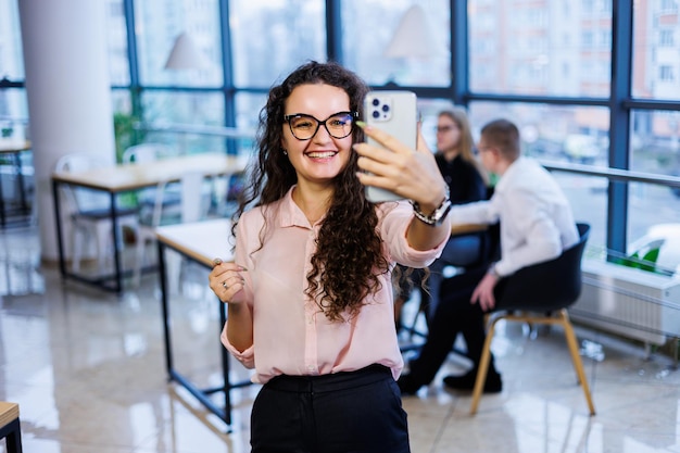 Jeune femme d'affaires intelligente et heureuse, vêtue de vêtements et de lunettes élégants, travaille au bureau, parle avec un client sur un smartphone, fait des gestes, sourit. Les employés travaillent en arrière-plan. Mise au point sélective