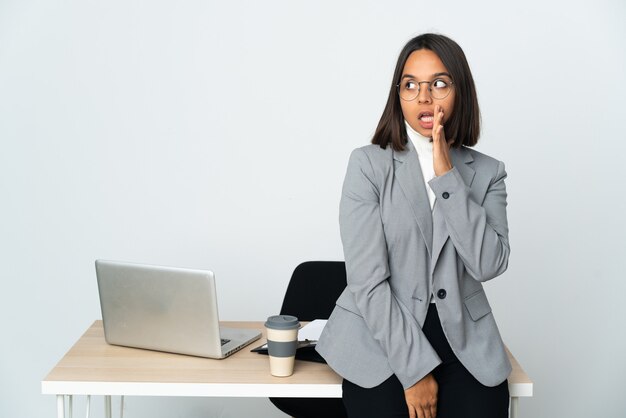 Jeune femme d'affaires latine travaillant dans un bureau isolé sur fond blanc chuchotant quelque chose avec un geste de surprise tout en regardant sur le côté