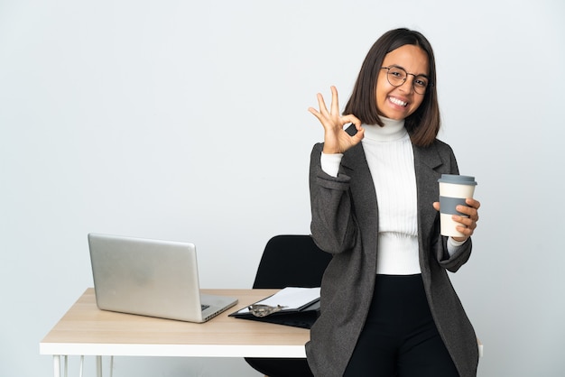 Jeune femme d'affaires latine travaillant dans un bureau isolé sur un mur blanc montrant signe ok avec les doigts