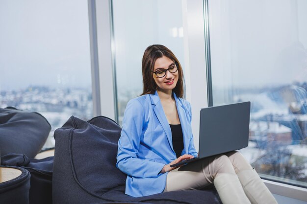 Jeune femme d'affaires à lunettes avec de longs cheveux noirs dans des vêtements décontractés souriant et regardant un ordinateur portable tout en parcourant des documents en ligne pendant le week-end dans l'espace de travail