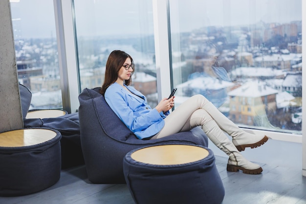 Jeune femme d'affaires à lunettes avec de longs cheveux noirs dans des vêtements décontractés souriant et regardant le téléphone naviguant sur le smartphone pendant une journée de congé dans l'espace de travail