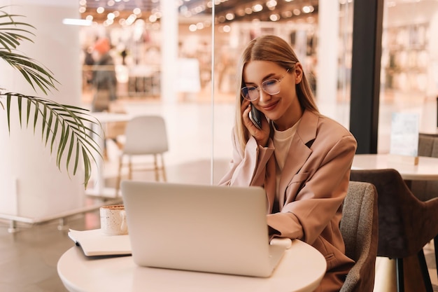Une jeune femme d'affaires avec des lunettes parle au téléphone et travaille en ligne à l'aide d'un ordinateur portable dans un café