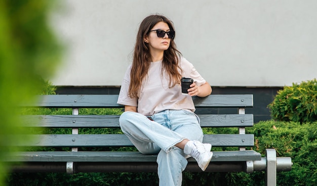 Une jeune femme d'affaires à lunettes de soleil est assise sur un banc avec une tasse de café