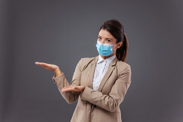 Jeune femme d'affaires avec masque facial debout et faisant des gestes avec les mains comme elle montre quelque chose.