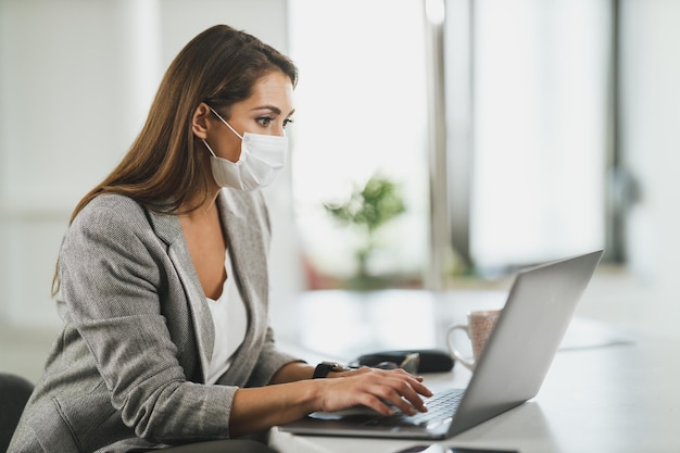 Jeune femme d'affaires avec masque de protection assise seule dans son bureau à domicile et travaillant sur un ordinateur portable pendant la pandémie du virus corona.