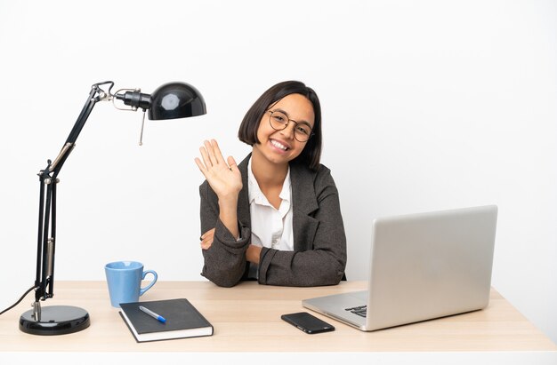 Jeune femme d'affaires mixte travaillant au bureau saluant avec la main avec une expression heureuse