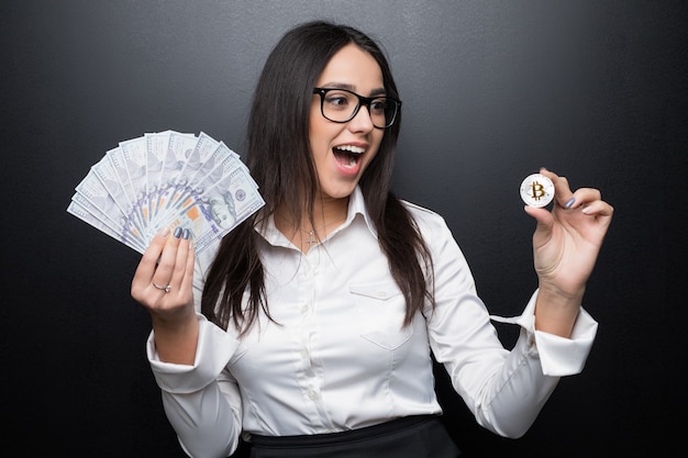 Jeune femme d'affaires moderne avec succès tenant un bitcoin d'or et un dollar en espèces isolé sur un mur noir