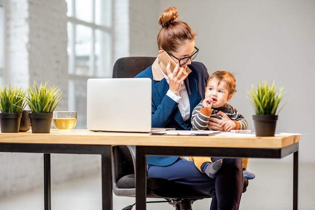 Jeune femme d'affaires multitâche vêtue du costume travaillant avec un ordinateur portable et des documents assis avec son bébé au bureau