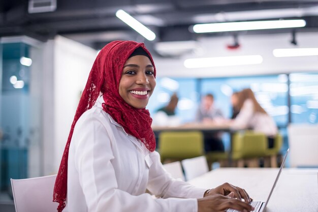 Jeune femme d'affaires musulmane noire moderne portant un hijab rouge, travaillant sur un ordinateur portable dans un bureau de démarrage. Diversité, concept multiracial