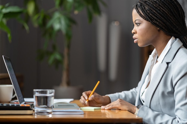 Jeune femme d'affaires noire concentrée sur l'écran d'un ordinateur portable en écrivant des notes