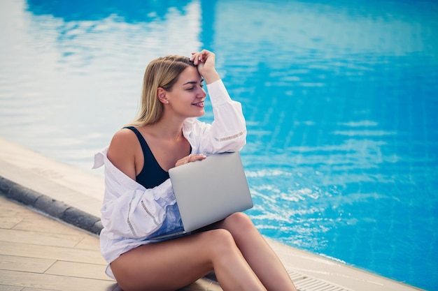 Jeune femme d'affaires avec un ordinateur portable assis au bord de la piscine Lieu de travail ouvert Concept de pigiste Achats en ligne Travail de vacances