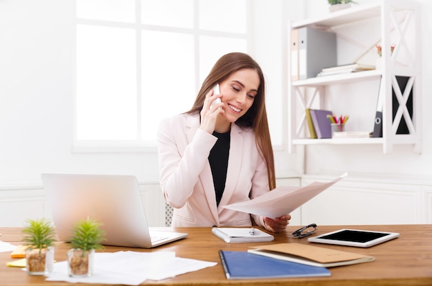 Jeune femme d'affaires parlant par téléphone avec des papiers, assise sur un lieu de travail de bureau moderne. Conseil aux entreprises.