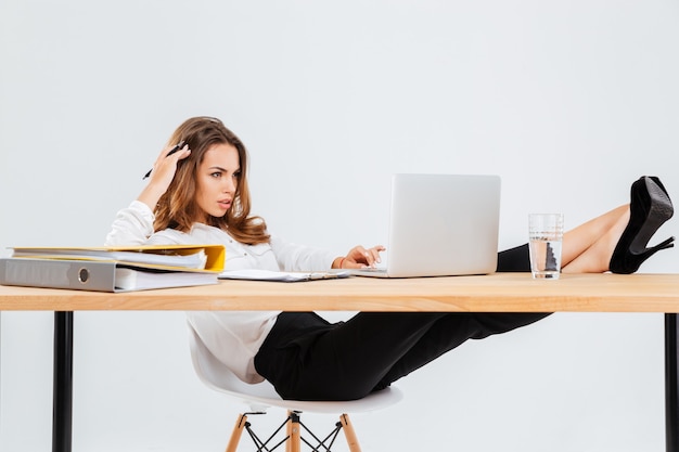 Jeune femme d'affaires pensive utilisant un ordinateur portable et pensant avec des jambes sur une table sur fond blanc