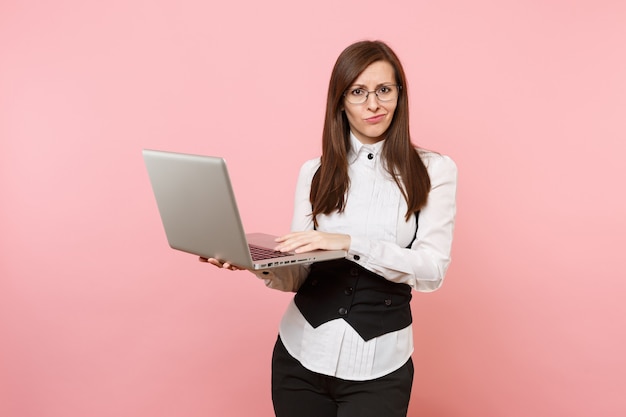 Jeune femme d'affaires perplexe en costume et lunettes travaillant dans un ordinateur portable isolé sur fond rose pastel. Dame patronne. Concept de richesse de carrière de réalisation. Copiez l'espace pour la publicité.