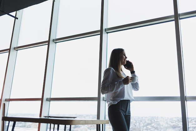 Jeune femme d'affaires portant des lunettes et tenant un téléphone Dans un bureau ou un centre d'affaires sur fond de grandes fenêtres d'un gratte-ciel Concept de travail et de carrière