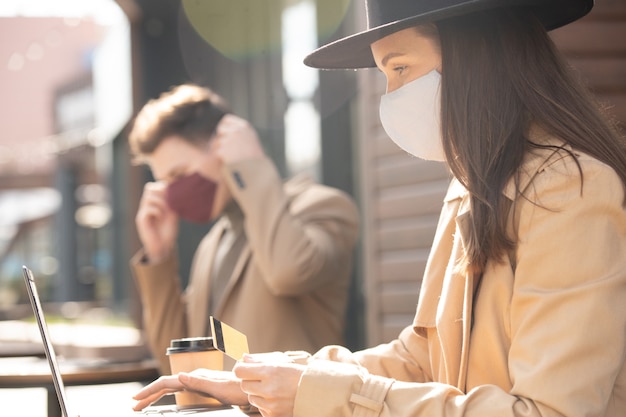 Jeune Femme D'affaires Portant Un Masque De Protection Tenant Une Carte De Crédit Tout En Faisant Des Achats En Ligne Devant Un Ordinateur Portable Et En Effectuant Le Paiement