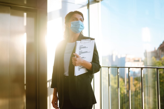 Une jeune femme d'affaires portant un masque de protection utilise un coude pour appuyer sur le bouton de l'ascenseur.