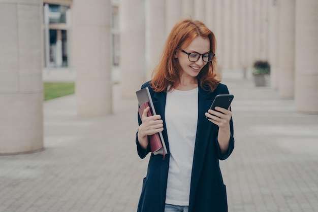 Jeune femme d'affaires positive aux cheveux roux en tenue élégante utilisant un téléphone portable, envoyant des SMS ou en vérifiant ses e-mails tout en se tenant à l'extérieur après avoir travaillé dur, tenant un ordinateur portable et un carnet de notes