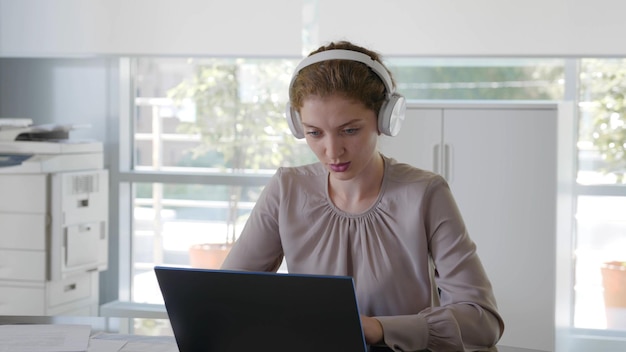 Jeune femme d'affaires positive dans les écouteurs tapant dans un ordinateur portable au bureau