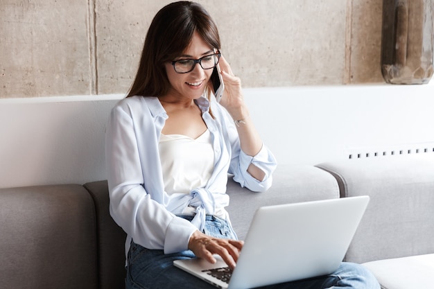 jeune femme d'affaires positive et joyeuse à l'intérieur à la maison sur un canapé dans le salon à l'aide d'un ordinateur portable parlant par téléphone portable.