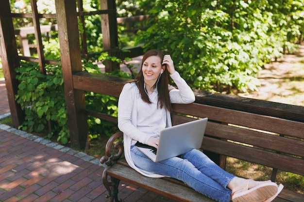 Jeune femme d'affaires prospère dans des vêtements légers et décontractés. Femme assise sur un banc travaillant sur un ordinateur portable moderne dans le parc de la ville dans la rue à l'extérieur sur la nature. Bureau mobile. Concept d'entreprise indépendant