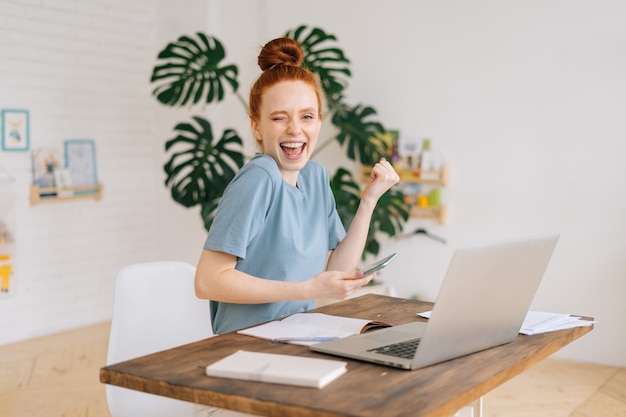 Une jeune femme d'affaires rousse heureuse et réussie utilise un téléphone portable et un ordinateur portable pour devenir bon