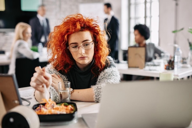 Jeune femme d'affaires rousse se sentant triste en mangeant de la salade et en lisant quelque chose sur un ordinateur au bureau