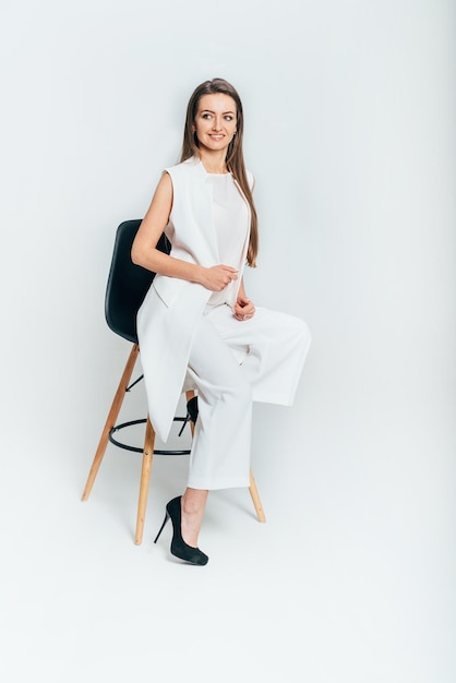 La jeune femme d'affaires s'assied sur une chaise dans le studio sur un fond blanc. Une femme en costume blanc et chaussures noires regarde sur le côté. Fermer