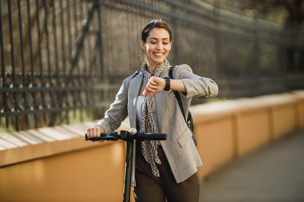 Une jeune femme d'affaires avec un scooter électrique va travailler et regarde une montre-bracelet.