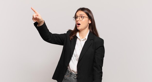 Jeune femme d'affaires se sentant choquée et surprise, pointant du doigt et regardant vers le haut avec admiration avec un regard étonné et bouche bée