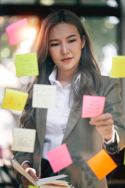 Jeune femme d'affaires séduisante souriante utilisant des notes autocollantes dans un mur de verre pour écrire un plan d'affaires stratégique pour le développement vers le succès