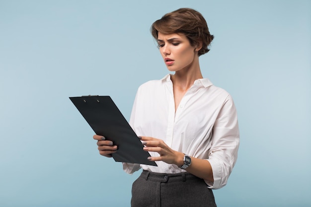 Jeune femme d'affaires sérieuse aux cheveux courts foncés en chemise blanche regardant tristement sur le dossier avec des documents sur fond bleu isolé