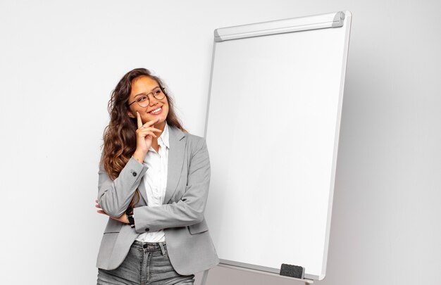 Jeune femme d'affaires souriant joyeusement et rêvassant ou doutant, regardant sur le côté