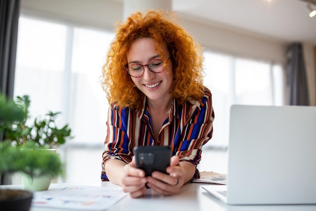 Jeune femme d'affaires souriante aux cheveux roux utilisant un smartphone près de lptop au bureau