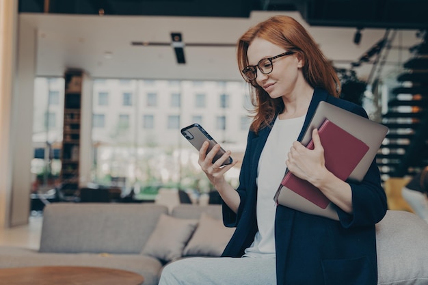 Jeune femme d'affaires souriante dans des verres avec ordinateur portable et agenda sous le bras en regardant un smartphone