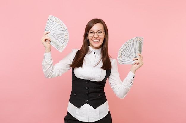 Jeune femme d'affaires souriante dans des verres tenant un paquet de dollars, de l'argent en espèces et écartant les mains isolées sur fond rose. Dame patronne. Réalisation carrière richesse. Copiez l'espace pour la publicité.