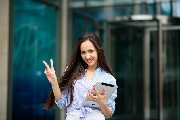 Jeune femme d'affaires souriante avec des documents administratifs et un ordinateur portable à la main prenant un selfie à l'extérieur par beau temps avec de vieux bâtiments en arrière-plan